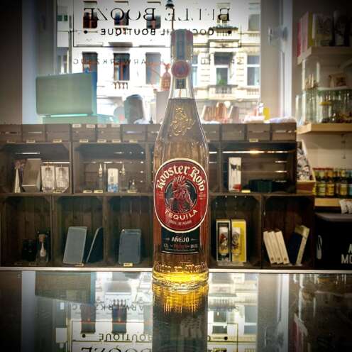 Produktbild Rooster Rojo Añejo Tequila Alt Attribut Rooster Rojo Añejo Tequila kaufen bei Belle Booze Tequila Glas mit goldener Farbe Alt Attribut Rooster Rojo Añejo gereifte Agave im Glas Flasche vor Agavenfeld Kulisse Alt Attribut Rooster Rojo Añejo Tequila aus Jalisco Mexiko