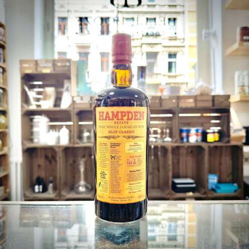 Produktbild Hampden HLCF Classic Rum Alt-Attribut: Hampden HLCF Classic Rum kaufen bei Belle Booze Glas mit HLCF Classic und tropischen Früchten Alt-Attribut: Hampden HLCF Classic Rum mit Ananas und Banane im Glas Flasche HLCF Classic vor Rumfässern Kulisse Alt-Attribut: Hampden HLCF Classic Rum – Jamaika High Ester Pot Still Rum